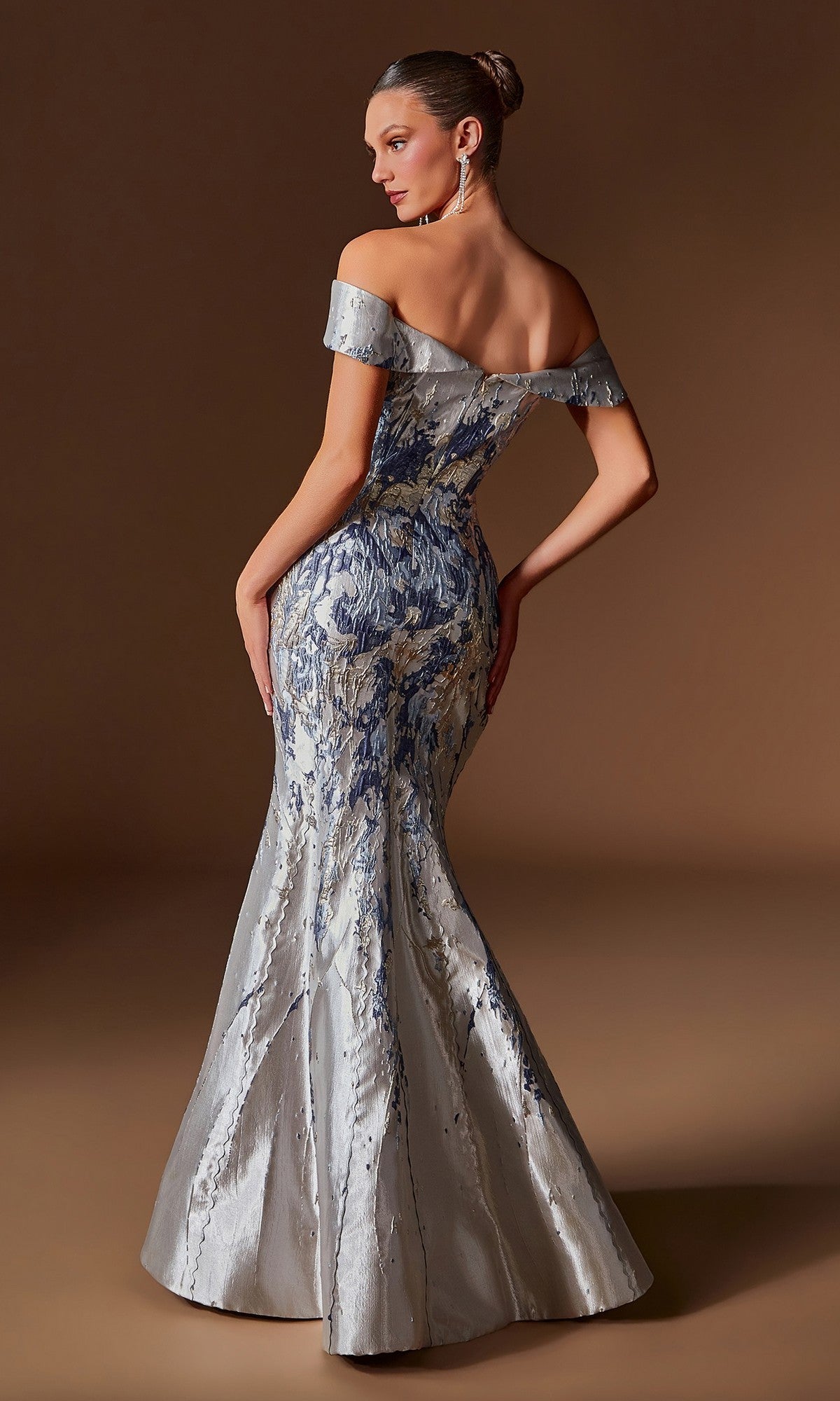 A woman models the Ladivine CD0107 long formal dress, featuring an off-the-shoulder silver and blue jacquard mermaid silhouette, as she poses against a plain brown background.